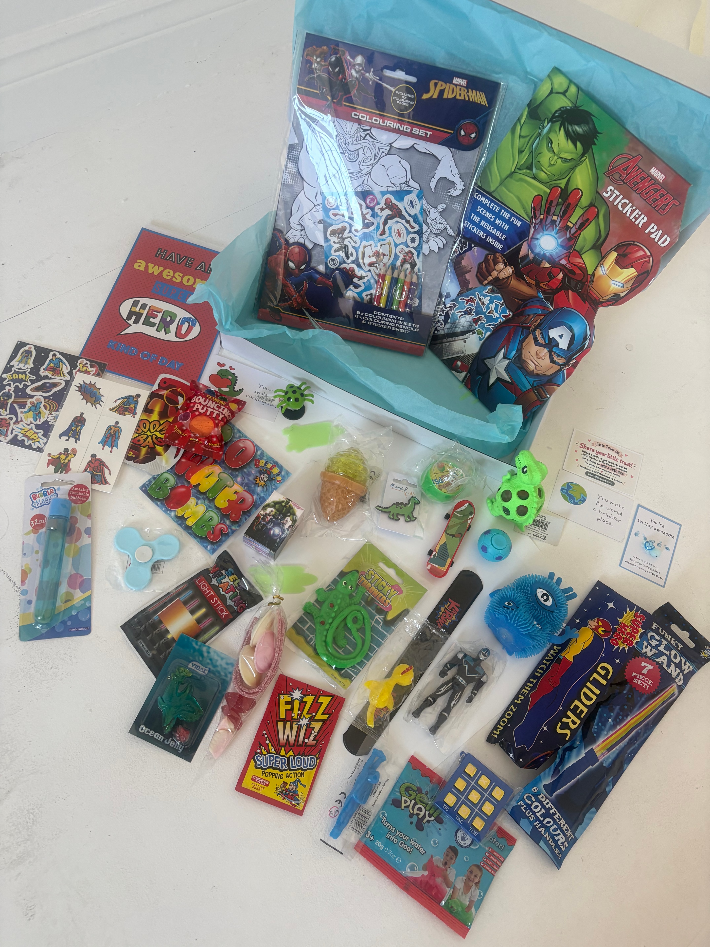Superhero themed children's gift box flat-lay showing Avengers sticker pad, Spider-Man colouring set, fidget toys and sweet treats